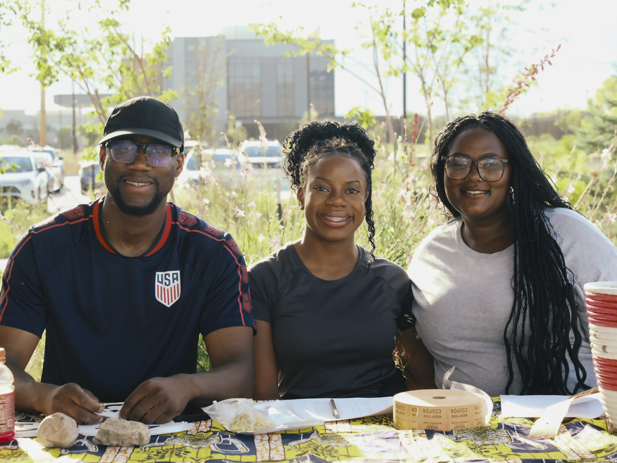 Three community members smiling together at a table during an outdoor gathering.