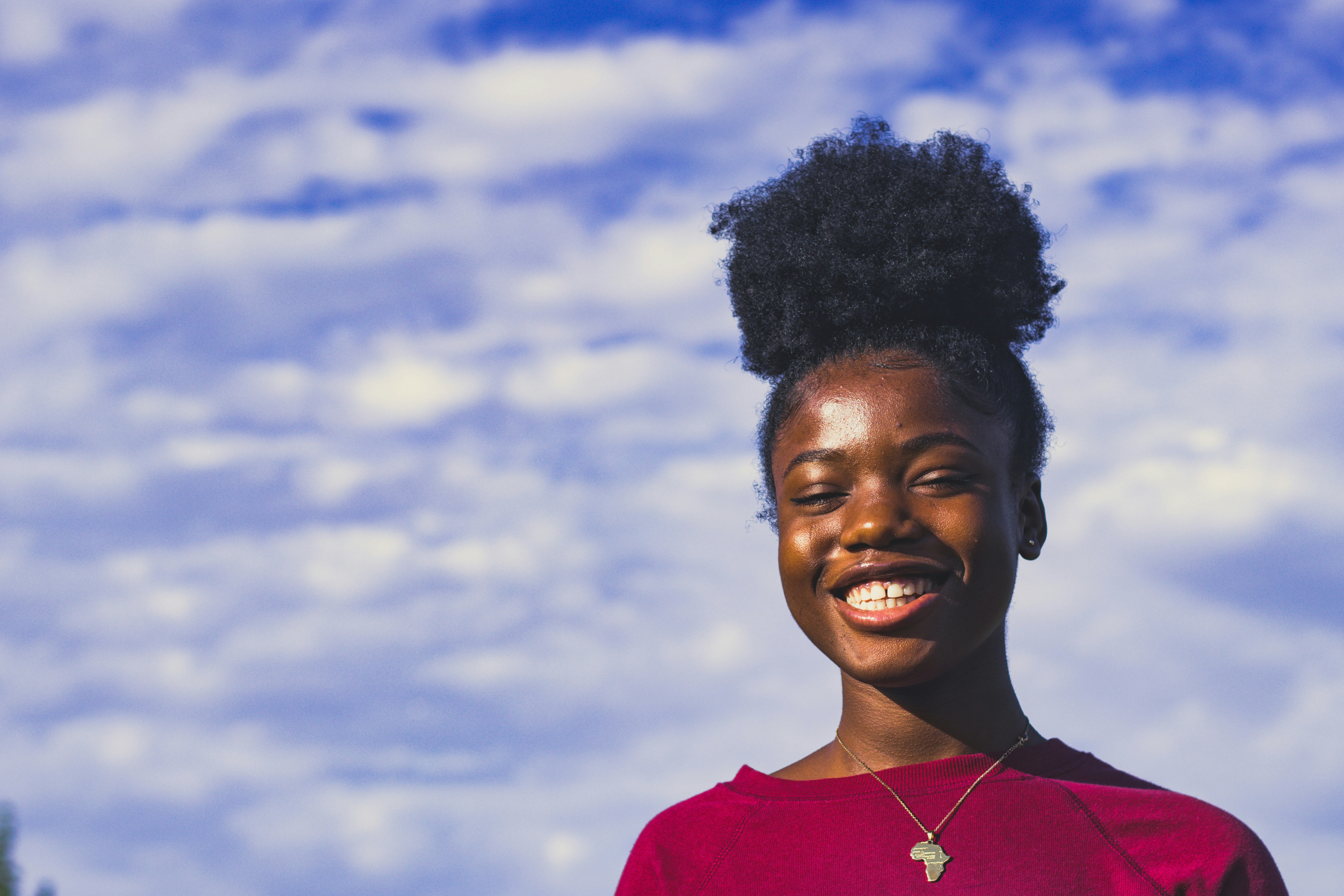 A smiling woman in a red sweater beneath a cloudy blue sky.